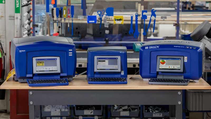 A Brady bbp85, s3100 and s3700 THT benchtop printer sitting on a table in a warehouse.